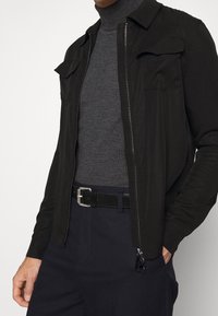 Veste légère noire avec fermeture éclair, deux poches poitrine et col, portée sur un col roulé gris foncé et associée à un pantalon bleu marine.