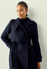 Navy wollen jas met een ceintuur in de taille, brede revers en zijzakken. Kenmerkend zijn de gestructureerde schouders en de gladde textuur.