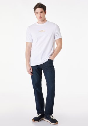Joven con camiseta blanca con el texto "Italy Amalfi Coast Naples", vaqueros azul oscuro y zapatillas negras, de pie sobre un fondo blanco.