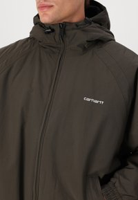 Veste vert olive foncé avec fermeture éclair à l'avant, capuche ajustable et poignets élastiques. Présente un logo Carhartt blanc sur la poitrine gauche.