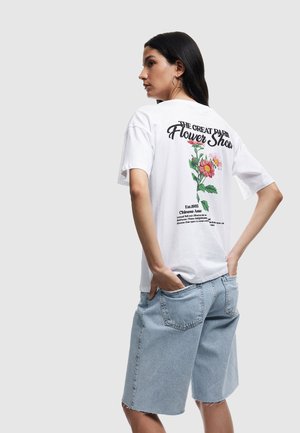 Hvid t-shirt med et blomsterdesign på ryggen, rund halsudskæring og korte ærmer. Sammen med lyseblå denimshorts med delvist frynset kant.