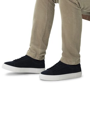 Sneakers in suede nera con una superficie liscia, suola in gomma bianca e punta arrotondata. Indossati con pantaloni in corduroy color beige chiaro.