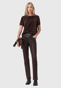 Maglione lavorato a maglia morbido di colore marrone scuro, maniche corte, abbinato a pantaloni di pelle marrone lucida. La modella tiene una cintura abbinata e indossa stivaletti scuri alla caviglia.