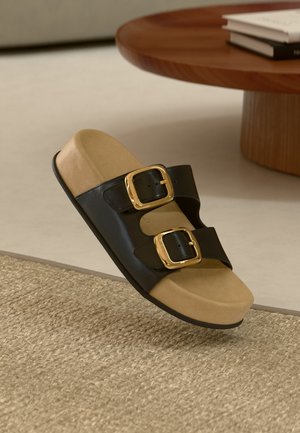 Sandale noire à glissière avec deux boucles dorées, un semelle beige texturée et une semelle lisse. Positionnée légèrement au-dessus d'un tapis tissé.