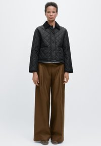 Zwarte gewatteerde jas met kraag en twee voorzakken, gecombineerd met brede bruine broek en grijze schoenen, tegen een effen achtergrond.