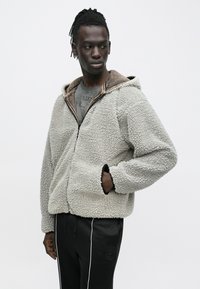 Junger Mann, der eine graue Sherpa-Fleecejacke mit Reißverschluss über einem grafischen T-Shirt und schwarzen Hosen mit weißen Seitenstreifen trägt, und zur Seite schaut.