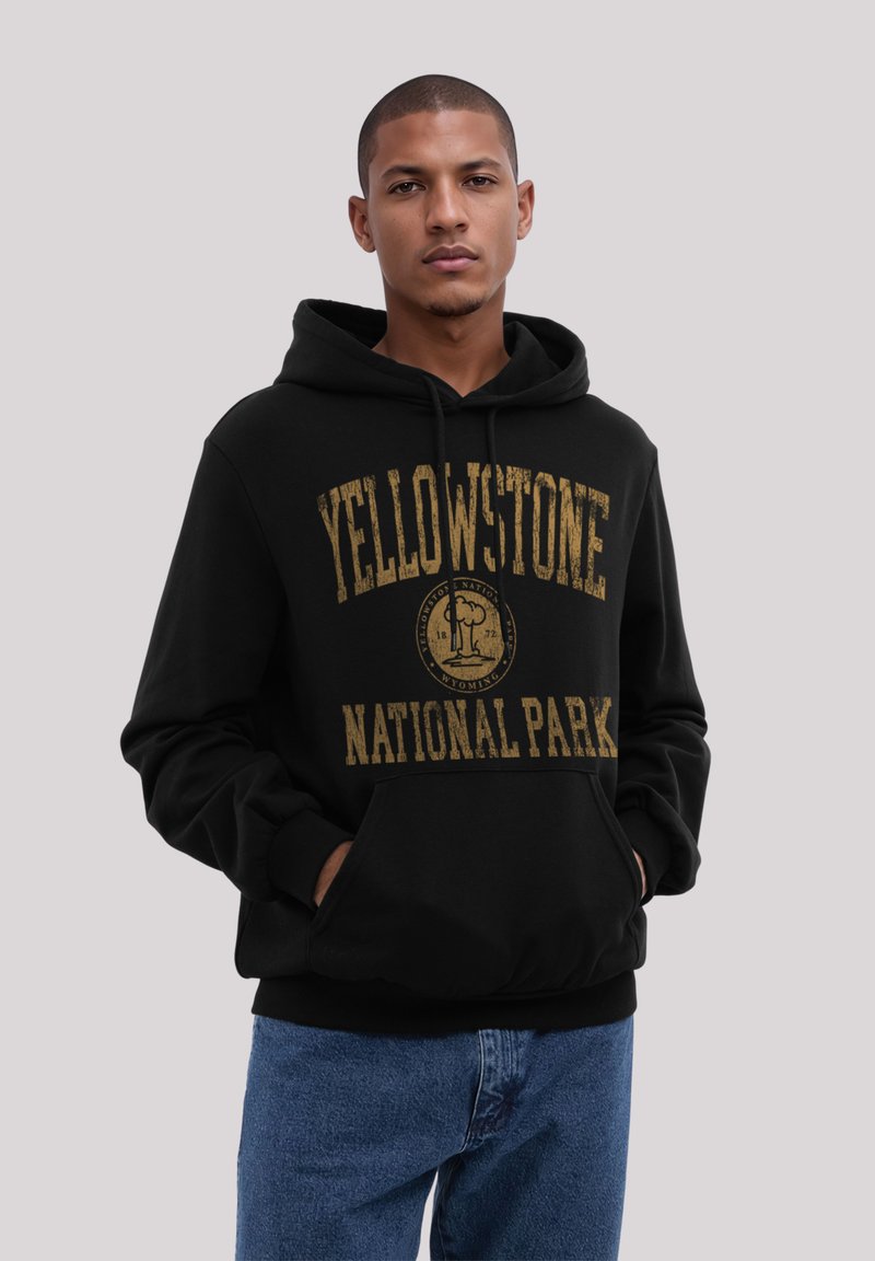Junger Mann mit kurzem Haar, der einen schwarzen Hoodie des Yellowstone-Nationalparks und eine blaue Jeans trägt, steht mit den Händen in den Taschen.