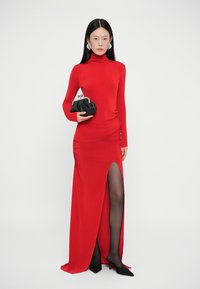 Lang rode jurk met een nauwsluitende col en een zijsplit, gemaakt van gladde stof. Model houdt een zwarte clutch vast en draagt zwarte hoge hakken.