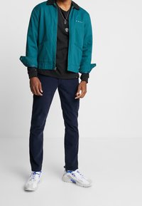 Turkos zip-up jacka med svart krage, svart långärmad tröja, mörkblå byxor och vita sneakers med grå detaljer.