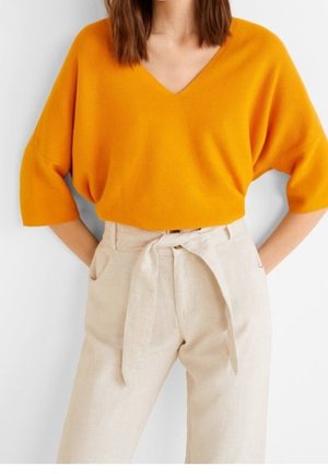 Mujer con un suéter naranja brillante de cuello en V y mangas hasta el codo, y pantalones beige de talle alto con cinturón atado.