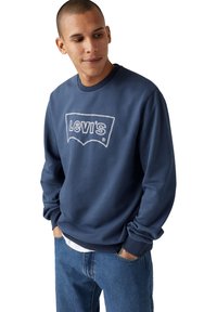 Donkerblauwe sweatshirt van zacht materiaal met een wit geborduurd Levi's-logo. Ontspannen pasvorm met ribgebreide manchetten en onderkant. Jeans eronder gedragen.