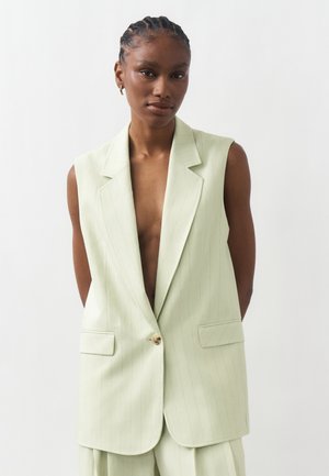 Naine põimitud juustega, kes kannab helerohelise triibulise varrukateta jopet üksiku nupuga ja sobivaid pükse, taustaks on lihtne taust.