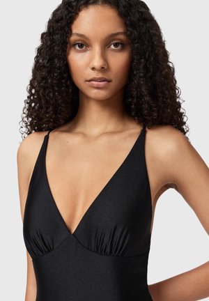 Nuori nainen, jolla on kiharat hiukset ja musta hihaton V-kauluksinen paita, yksinkertaisen vaalean taustan edessä.