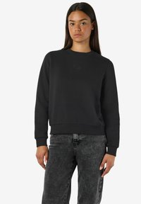 Joven mujer con cabello largo y oscuro, vestida con un sudadera negra lisa y jeans negros desgastados, de pie frente a un fondo blanco.