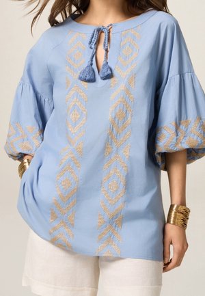 Lichtblauwe katoenen blouse met gouden borduursels in diamantpatroon, een sleutelgat-halslijn en korte puffmouwen. Gecombineerd met witte short.