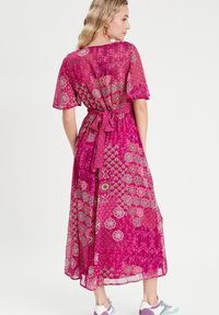 Robe longue rose avec un motif floral et géométrique, manches courtes, ceinture à nouer à la taille et texture fluide et transparente.