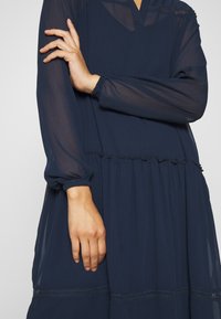 Personne en robe longue bleu marine à manches longues avec tissu transparent aux manches, un bras croisé sur la poitrine, l'autre pendant.