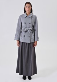 Cappotto grigio doppiopetto con cintura, dotato di ampie rever e tasche laterali, abbinato a una lunga gonna grigia e stivali neri alla caviglia.