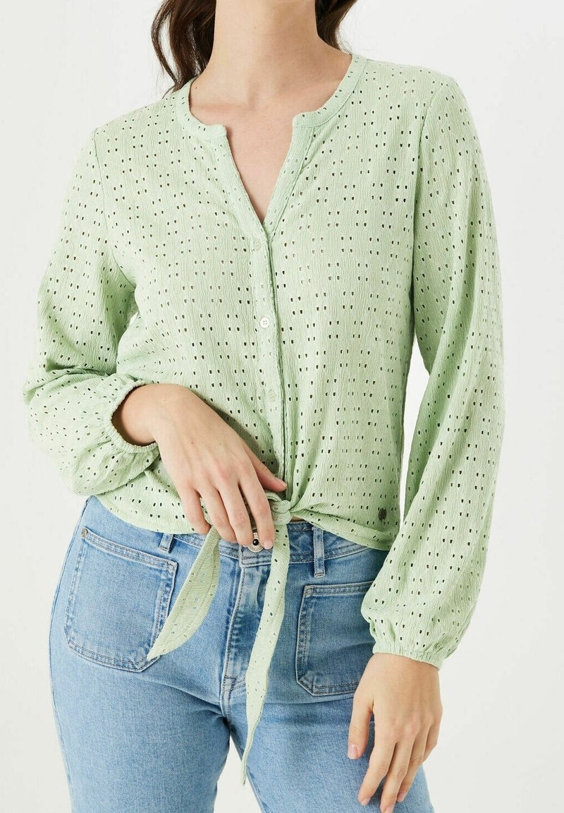 Kobieta w jasnozielonej bluzce z ażurowym wzorem, z długim rękawem, zawiązanej w talii, oraz w jasnoniebieskich dżinsach, stojąca na jednolitym tle.