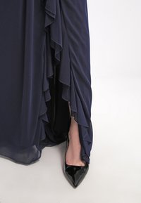 Pied d'une femme dans une chaussure noire en cuir verni à bout pointu visible sous une robe bleu foncé à volants sur un sol blanc.