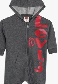 Szary, dresowy kombinezon z kapturem, wykonany z miękkiej tkaniny, z pełnym zamkiem błyskawicznym oraz dużym czerwonym logo "Levi's" z przodu. Posiada boczne kieszenie.