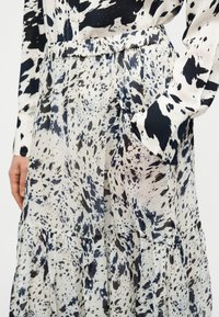 Μαύρο και λευκό φόρεμα με μοτίβο, φτιαγμένο από ελαφρύ ύφασμα, με σχεδιασμό πολλαπλών επιπέδων, σφηκοειδή μέση και μακριά μανίκια με λεπτομέρειες κουμπιών.