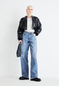 Veste bombardier en cuir noir avec des poignets à côtes, chemise blanche ajustée, jean clair à jambes larges, et bottes noires épaisses, tenant un sac sombre.