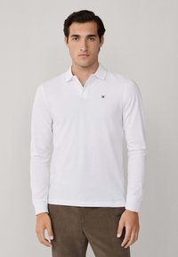 Mann trägt ein weißes Langarm-Polohemd mit einem kleinen gestickten Logo auf der Brust, kombiniert mit braunen Hosen, steht vor einem schlichten Hintergrund.