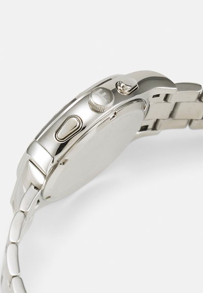 Strieborné kovové náramkové hodinky z bočného pohľadu zobrazujúce dva tlačidlá, jednu okrúhlu korunku s písmenom F a prepojený kovový náramok na bielom pozadí.