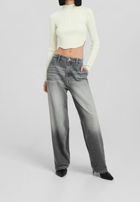 Kvinna i högmidjade, urtvättade svarta jeans, en kort creamfärgad ribbad långärmad tröja och svarta spetsiga klackar, stående med handen i fickan.