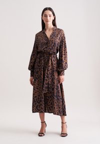 Abito a stampa leopardata con profondo scollo a V, silhouette fluida, maniche lunghe e cintura da annodare, realizzato in un tessuto liscio nelle tonalità di marrone e nero.