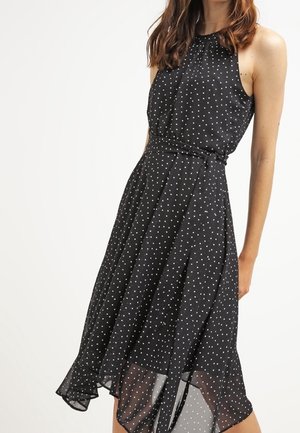 Frau trägt ein ärmelloses schwarzes Kleid mit weißen Punkten, tailliertem Schnitt und einem fließenden, asymmetrischen Saum vor einfachem Hintergrund.