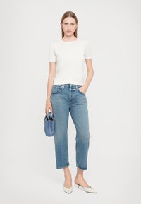 T-shirt bianco a maniche corte, jeans azzurri a vita alta, scarpe bianche con punta affusolata, che tiene in mano una piccola borsa blu, in piedi su uno sfondo neutro.