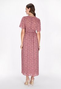 Maxi-jurk van roze stof met een bloemenpatroon. Heeft korte mouwen, een gesmokte taille en een vloeiende zoom. Draagt met beige hoge hakken.