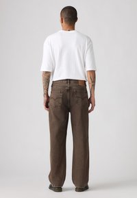Vaqueros de pierna ancha marrones con un parche de cuero en la parte posterior, costuras visibles y dos bolsillos traseros. Combinados con una camisa blanca de manga corta.