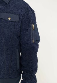 Granatowa bluza polarowa z teksturowanym, puszystym wykończeniem, kieszenią na ramieniu zapinaną na zamek błyskawiczny oraz mosiężnymi guzikami. Posiada dopasowany pas.