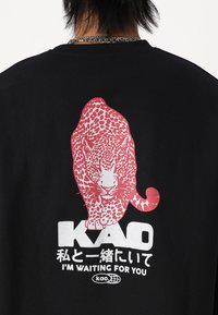 Svart t-skjorte med et stort rødt leopardgrafikk med detaljerte flekker, og hvit tekst som leser "KAO" og "JEG VENTER PÅ DEG" under.