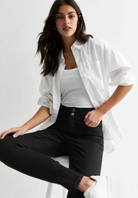 Chemise blanche à boutons sur un débardeur à côtes ajusté, associée à un jean noir taille haute et des baskets blanches, illustrant un look décontracté.