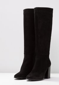 Bottes noires en daim montantes avec un talon carré, bout pointu et texture lisse. Le design présente un dessus sans couture et sans matériel visible.