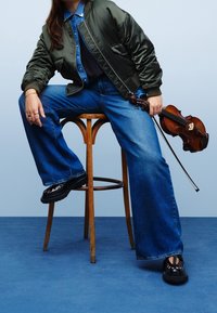 Personne portant une veste bomber verte et un jean à jambes larges, assise sur un tabouret en bois, tenant un violon et un archet devant un fond bleu clair.