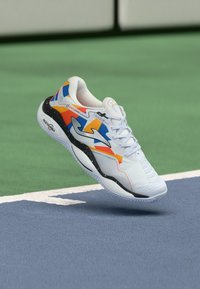 White athletic shoe with multicolored accents in orange, blue, and black. Features textured upper and distinctive sole design for grip.