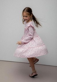 Młoda dziewczyna wirująca w różowej kwiecistej sukience z falbankami, w skarpetkach i różowych butach, uśmiechająca się na szarym tle.