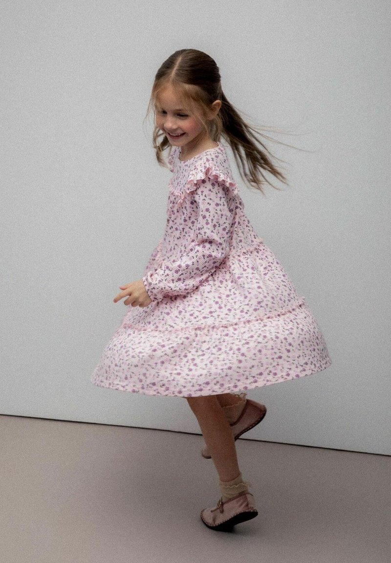 Młoda dziewczyna wirująca w różowej kwiecistej sukience z falbankami, w skarpetkach i różowych butach, uśmiechająca się na szarym tle.