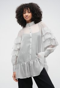 Vaaleanharmaa läpikuultava pusero röyhelövarusteilla ja kaksikerroksisella helmalla, jossa on korkea kaulus ja etupuolen nappikiinnitys. Sileä rakenne.