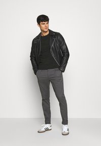 Chaqueta de cuero negra de motorista sobre un suéter oscuro, combinada con pantalones grises y zapatillas blancas con rayas negras. Figura masculina de pie de manera casual.