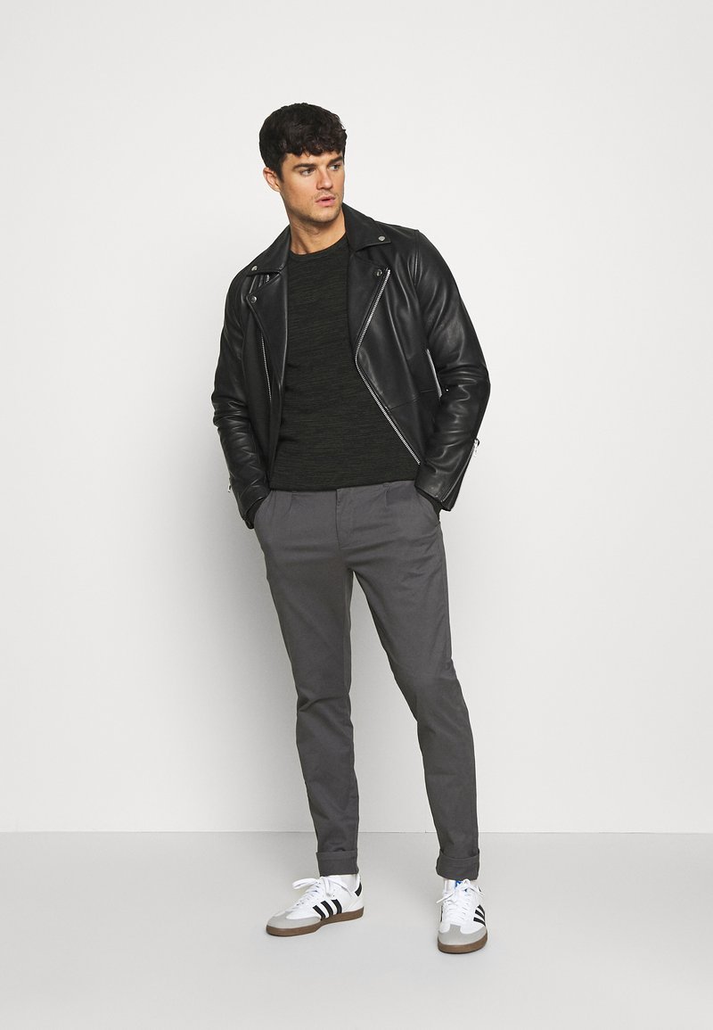 Chaqueta de cuero negra de motorista sobre un suéter oscuro, combinada con pantalones grises y zapatillas blancas con rayas negras. Figura masculina de pie de manera casual.