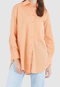 Chemise boutonnée en coton orange avec un col, des manches longues et une poche poitrine ; présente une coupe décontractée et une texture lisse.