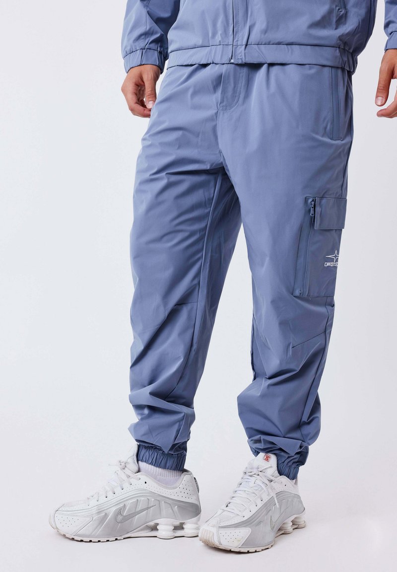 Man in blauwe cargo joggerpants met elastische boorden en witte sportschoenen, staand voor een effen lichte achtergrond.