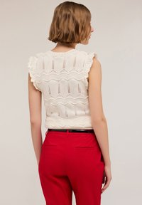 Top bianco lavorato a maglia con maniche arricciate, un motivo testurizzato con onde e linee verticali, indossato con pantaloni rossi su misura.