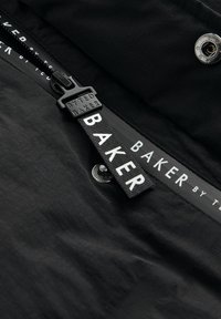 Tissu noir avec une texture lisse, comportant un tirette de fermeture éclair étiquetée "BYTED BAKER" et un bouton-pression en métal ; marque visible le long du bord.